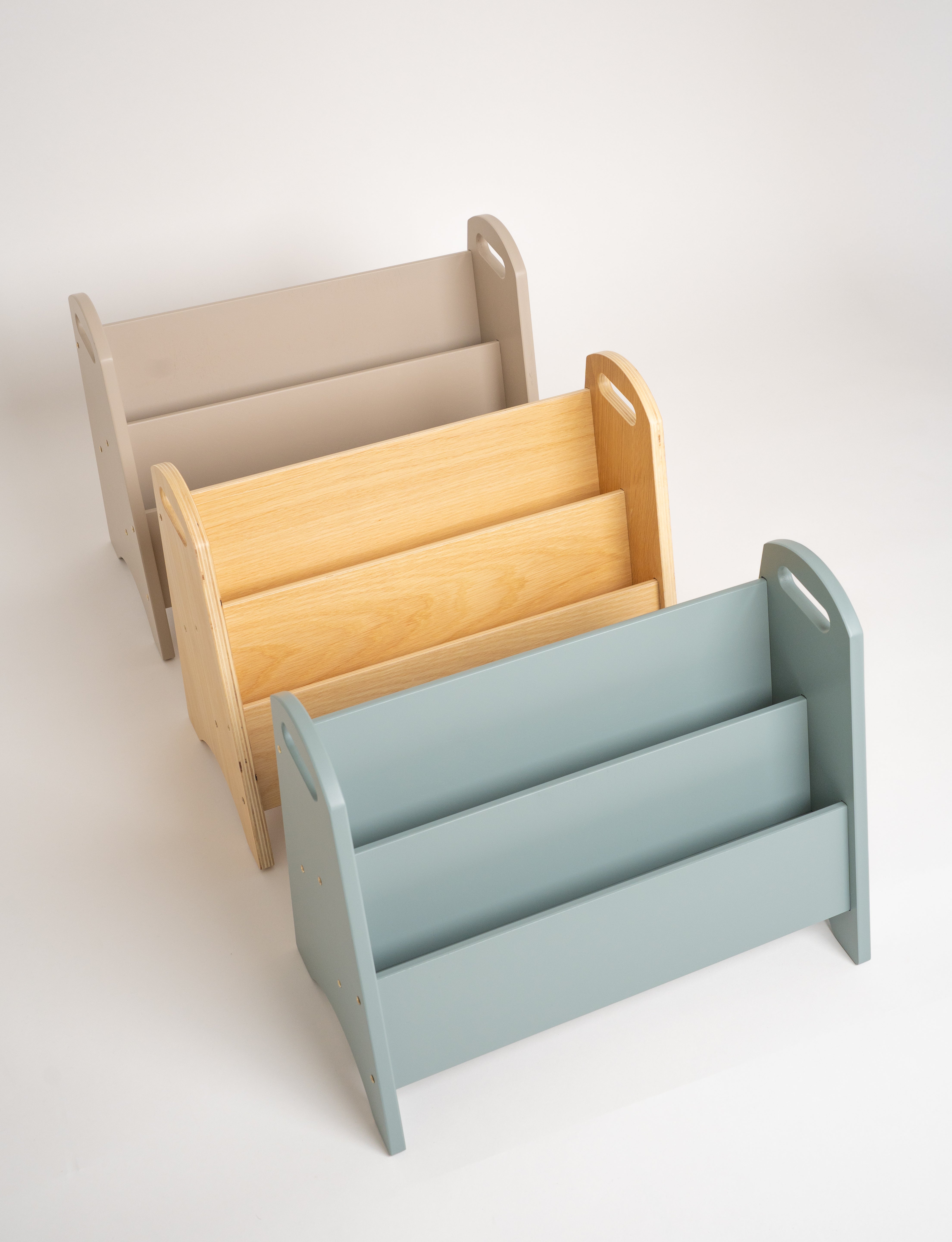 Three wooden magazine racks in different colors: beige, natural wood, and light blue, arranged diagonally on a white background.