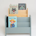 A light blue children's book organizer with three tiers, holding several colorful children's books, set against a plain light gray background.