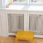 Curry stool in front of a white radiator with a window in the background