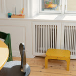 Children's room with a curry stool, green chair, and wooden table near a window.