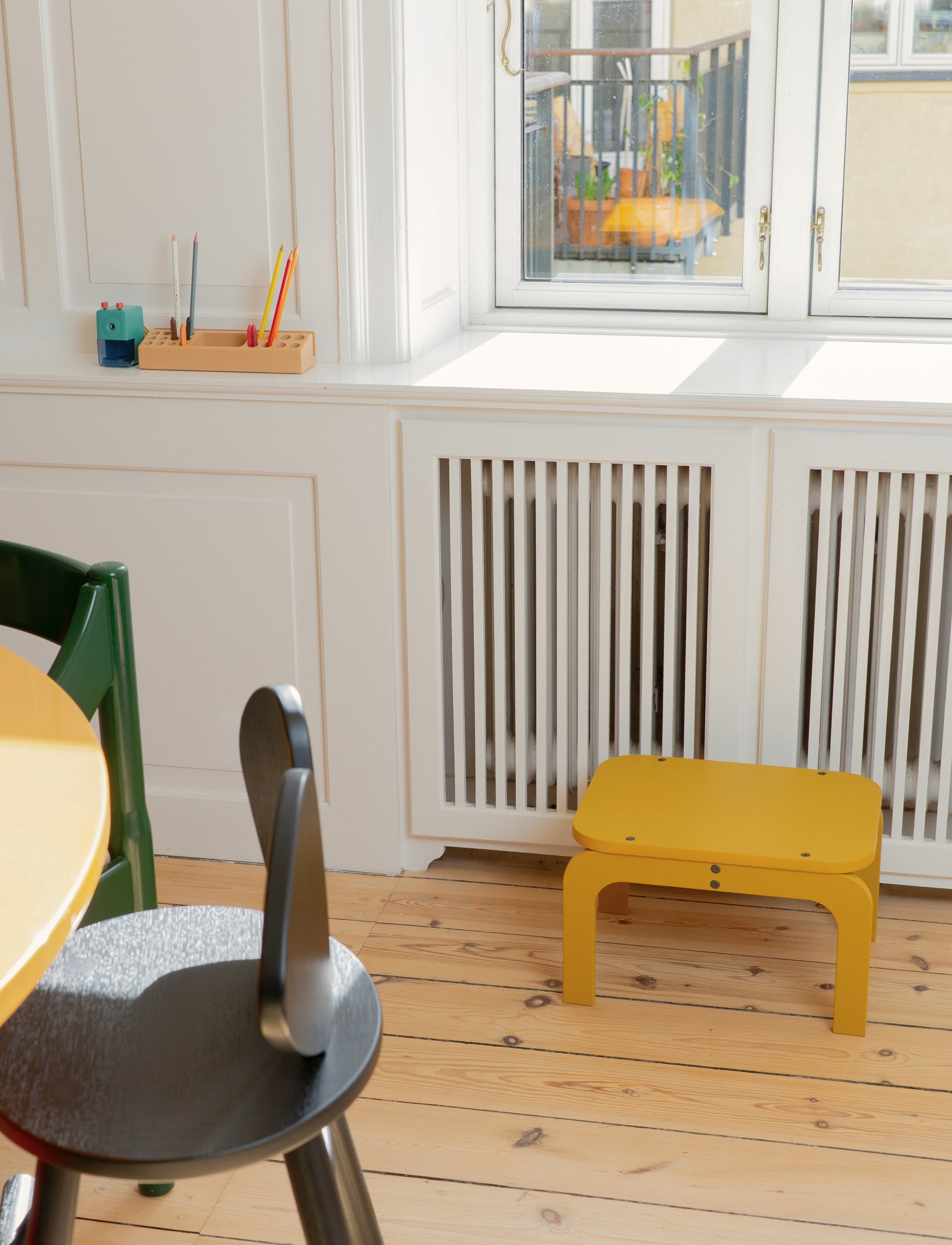Children's room with a curry stool, green chair, and wooden table near a window.