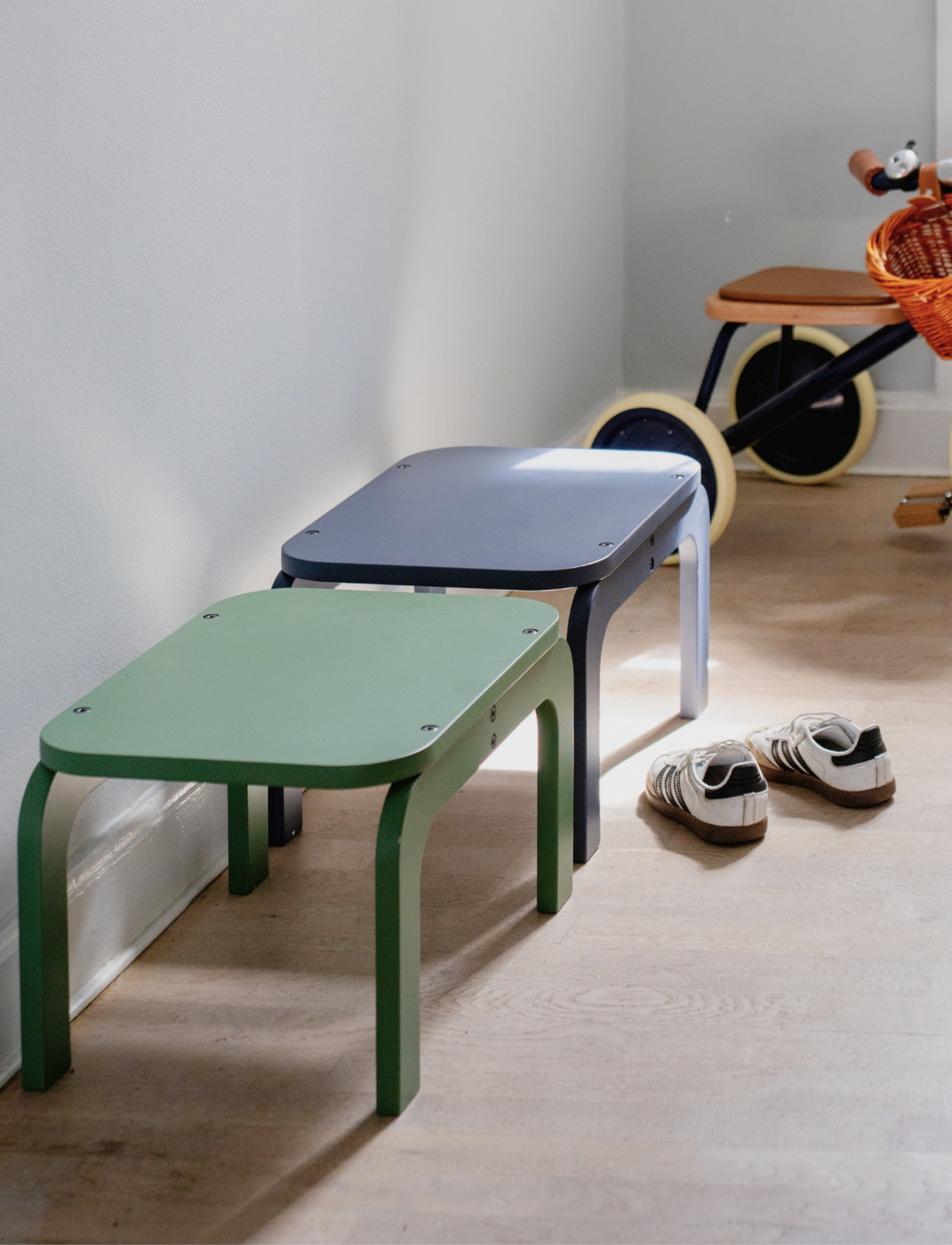 Two children's stools in green and blue on a wooden floor with a bicycle in the background.