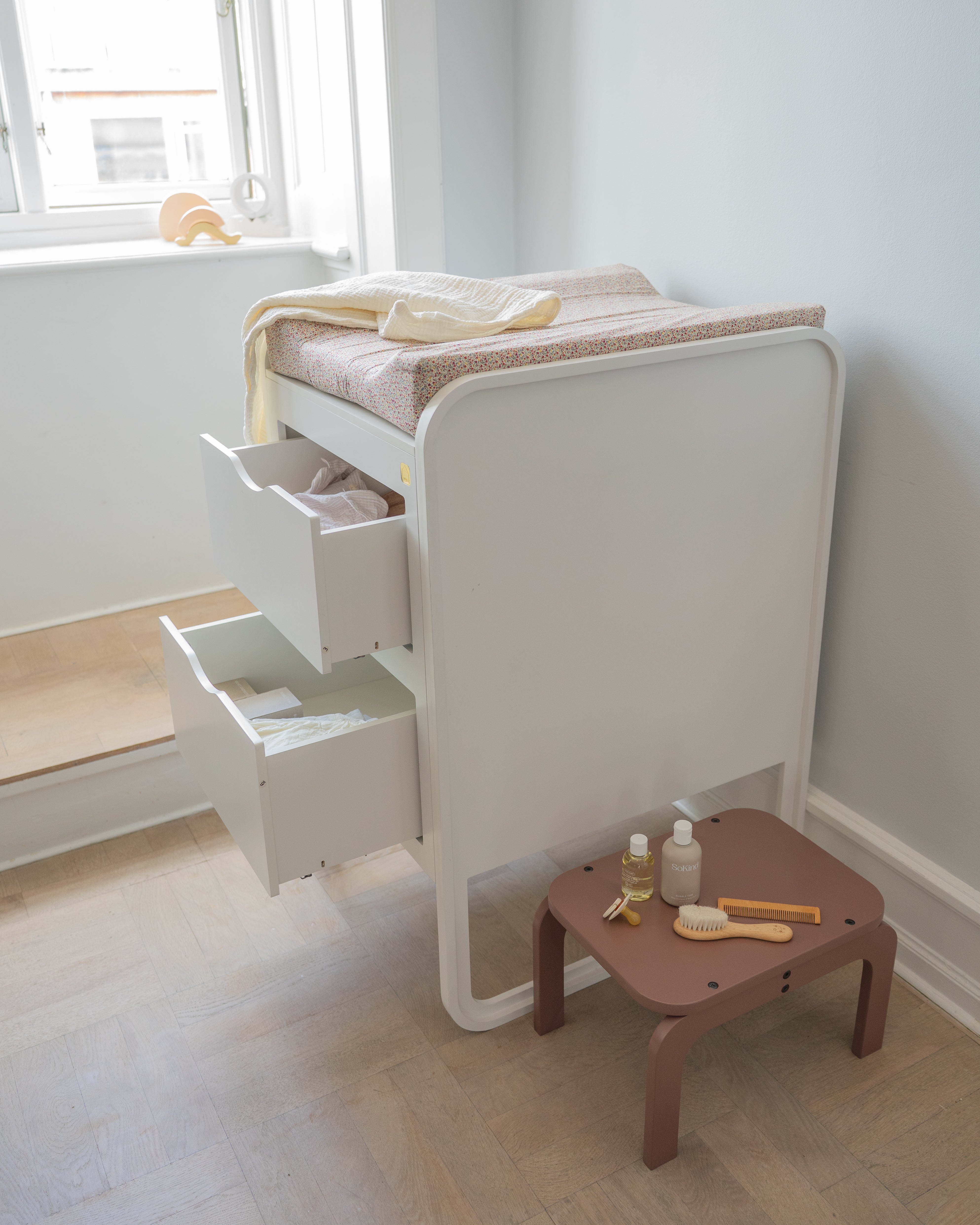 White changing table with drawers and a small brown table with baby care items in a room.