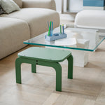 Green stool in front of a glass coffee table with stationery on a wooden floor.