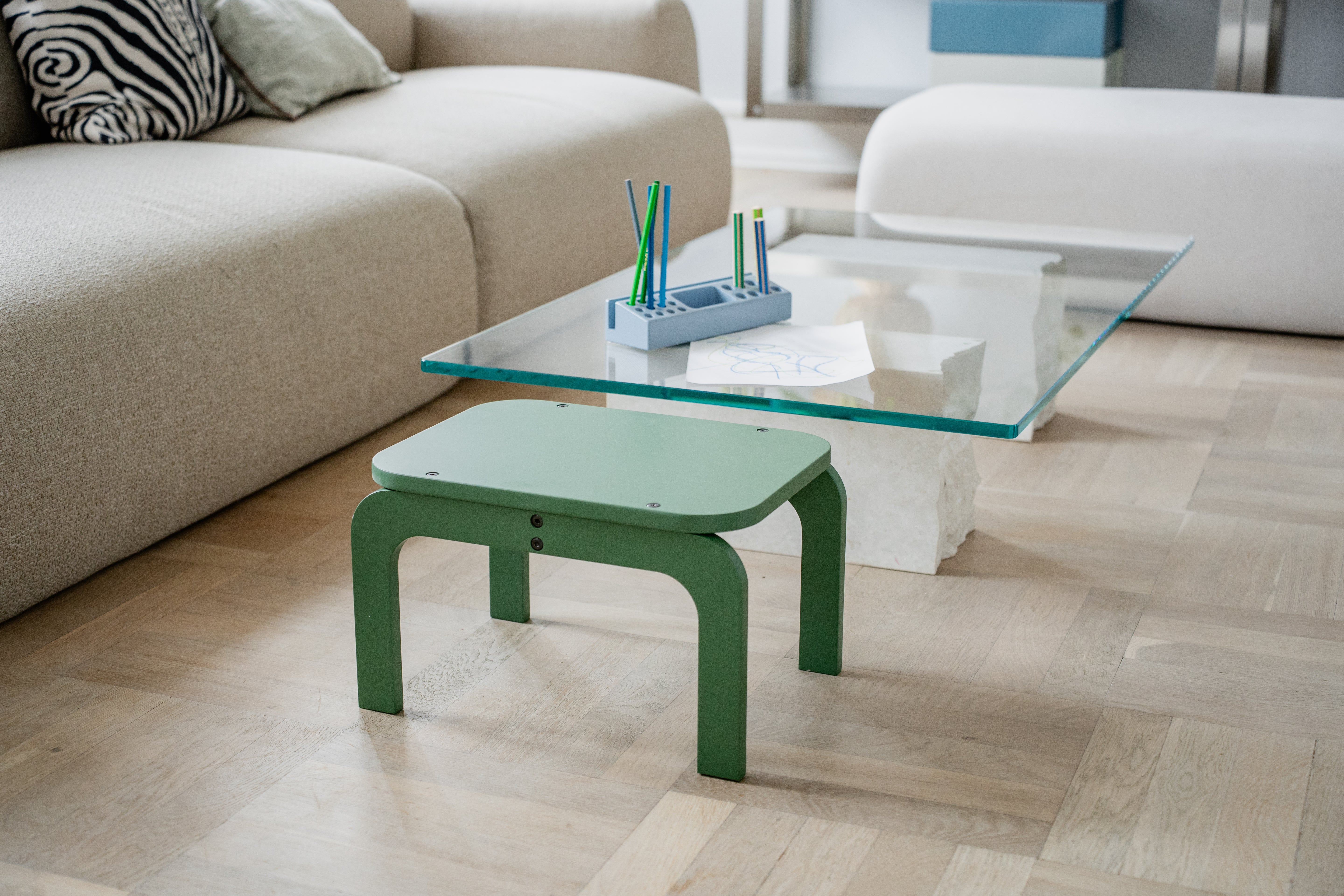 Green stool in front of a glass coffee table with stationery on a wooden floor.