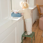 Child standing on a green step stool reaching into a blue box on a white surface.