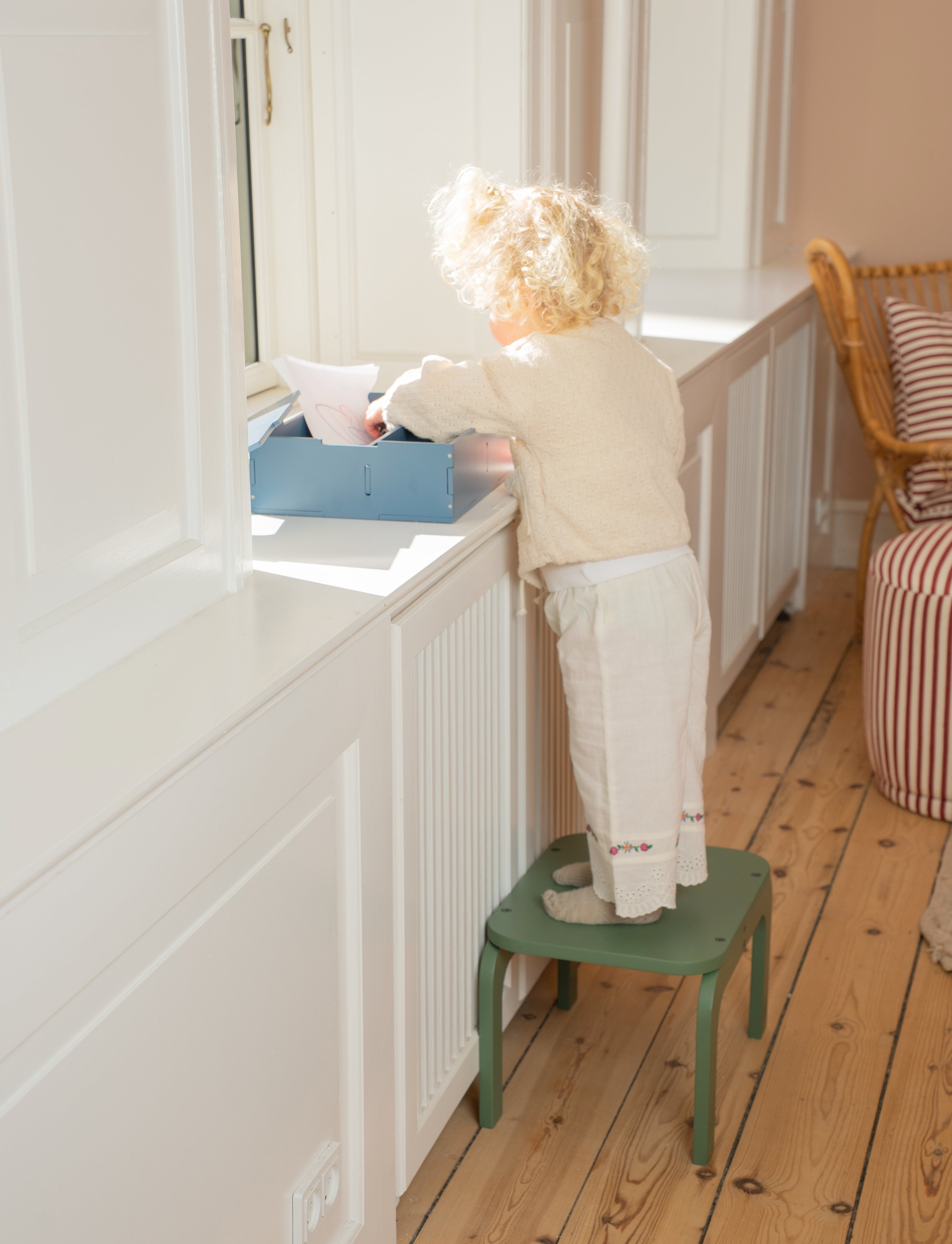 Child standing on a green step stool reaching into a blue box on a white surface.