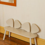 A minimalist children's bench with a curved cream-colored seat and four wing-like backrest panels, set against a striped wallpaper wall with a wooden floor.