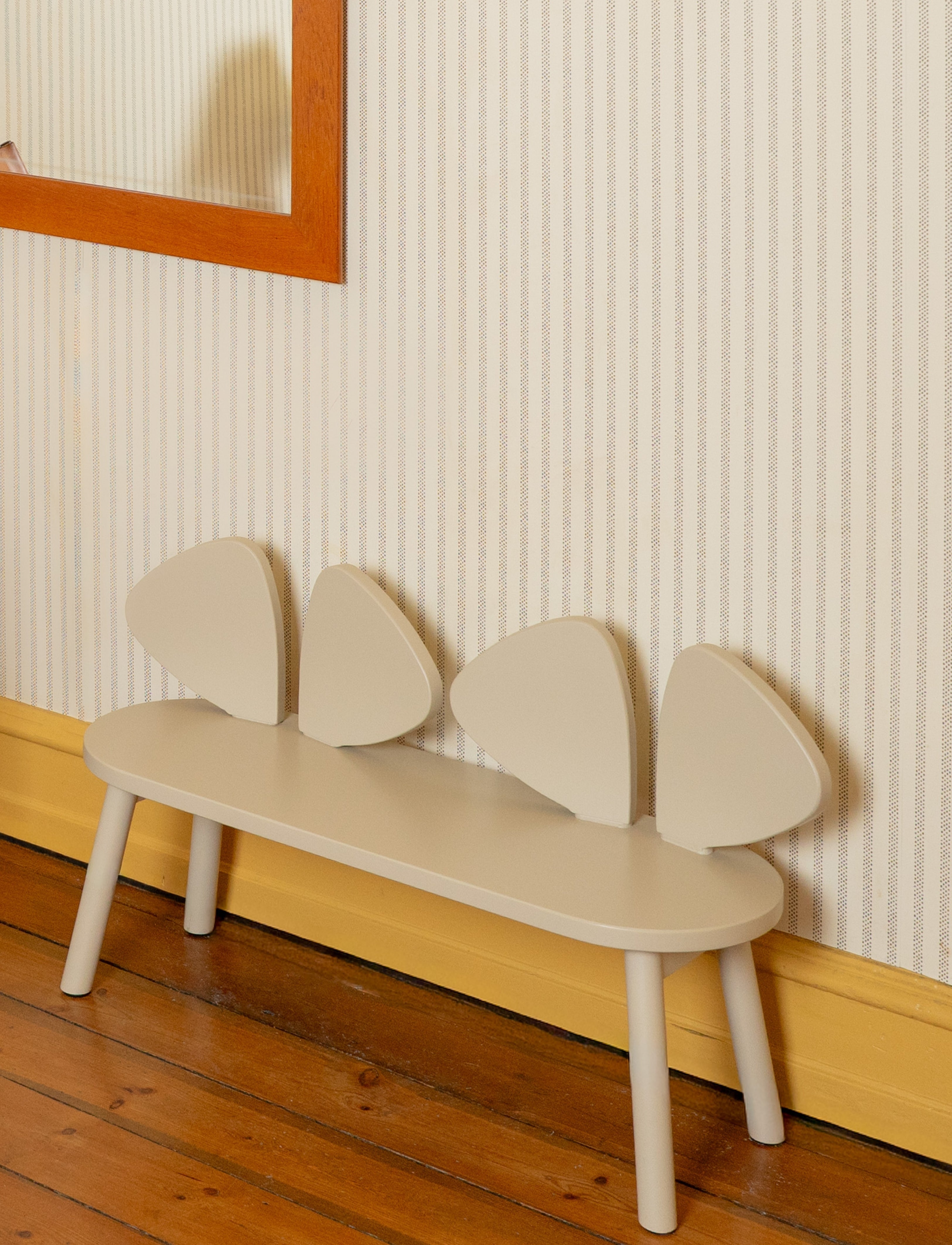 A minimalist children's bench with a curved cream-colored seat and four wing-like backrest panels, set against a striped wallpaper wall with a wooden floor.