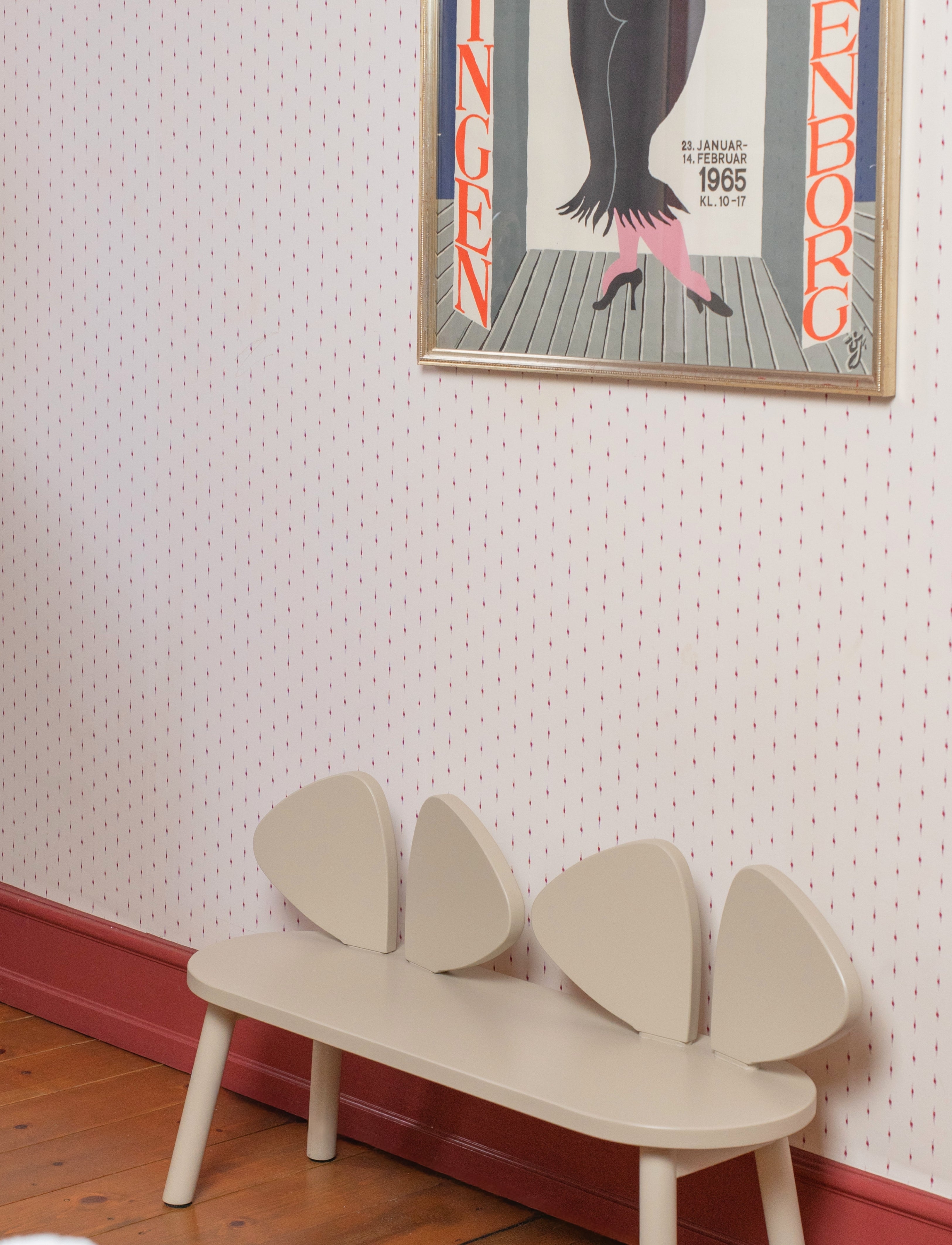A cream-colored wooden bench with three curved backrests, placed against a patterned wallpaper wall with a framed art print above it.