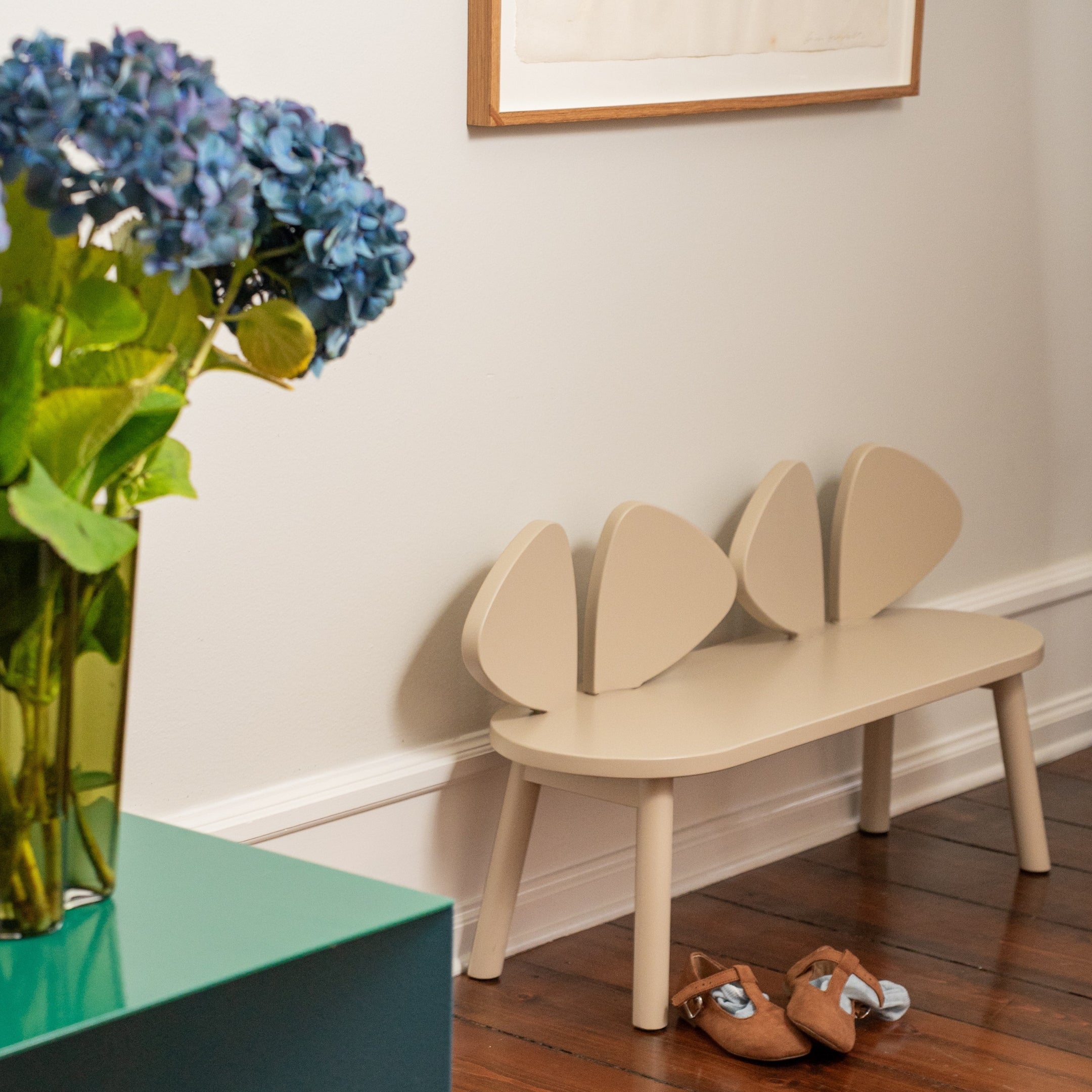 Beige children's bench with unique leaf design on a wooden floor, next to a vase of blue flowers.