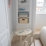 A small children's wooden table and matching chair in a bright, neutral nursery or playroom corner with built-in shelves, a window, and light wood flooring.