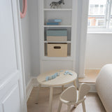 A small children's wooden table and matching chair in a bright, neutral nursery or playroom corner with built-in shelves, a window, and light wood flooring.