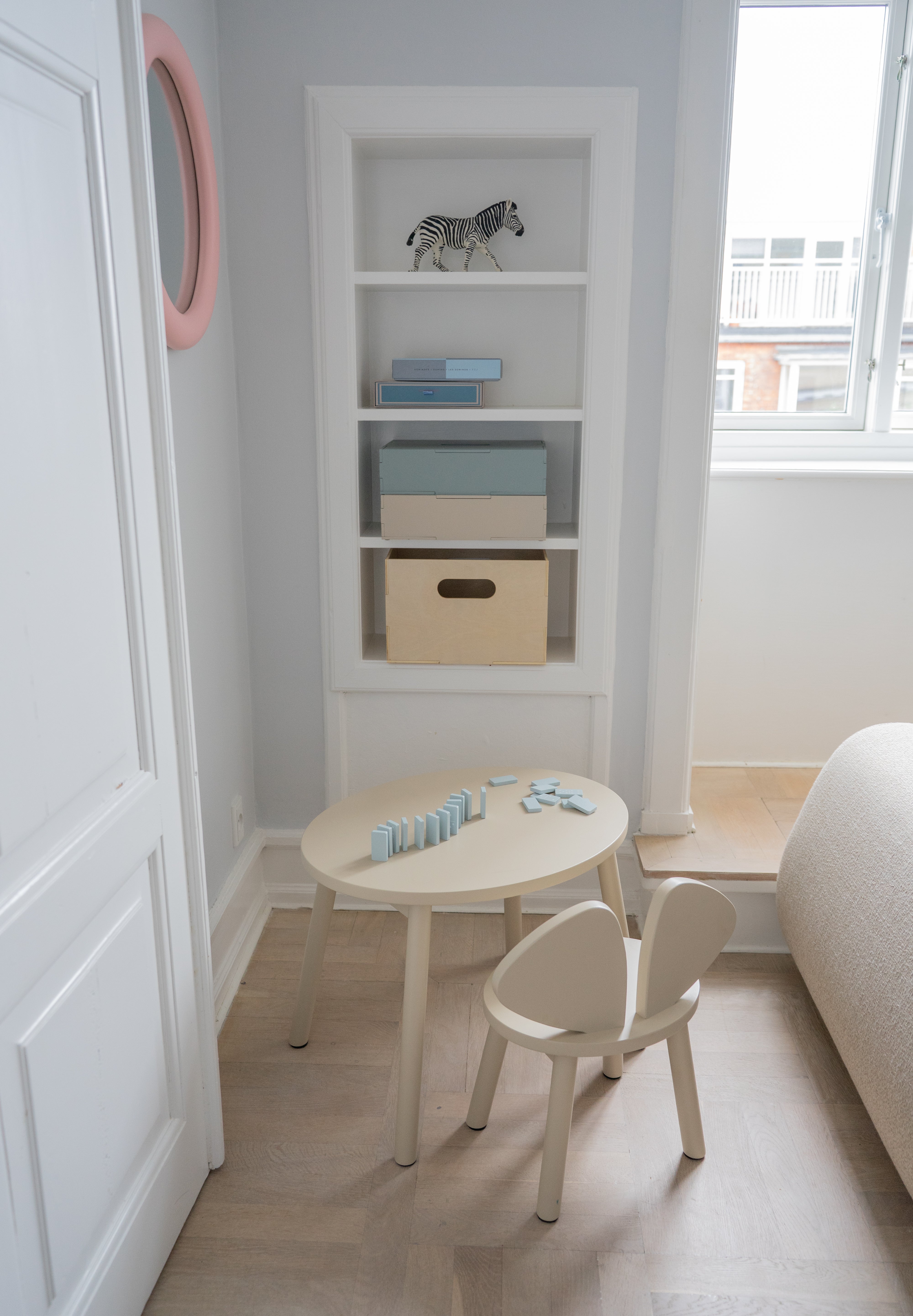 A small children's wooden table and matching chair in a bright, neutral nursery or playroom corner with built-in shelves, a window, and light wood flooring.