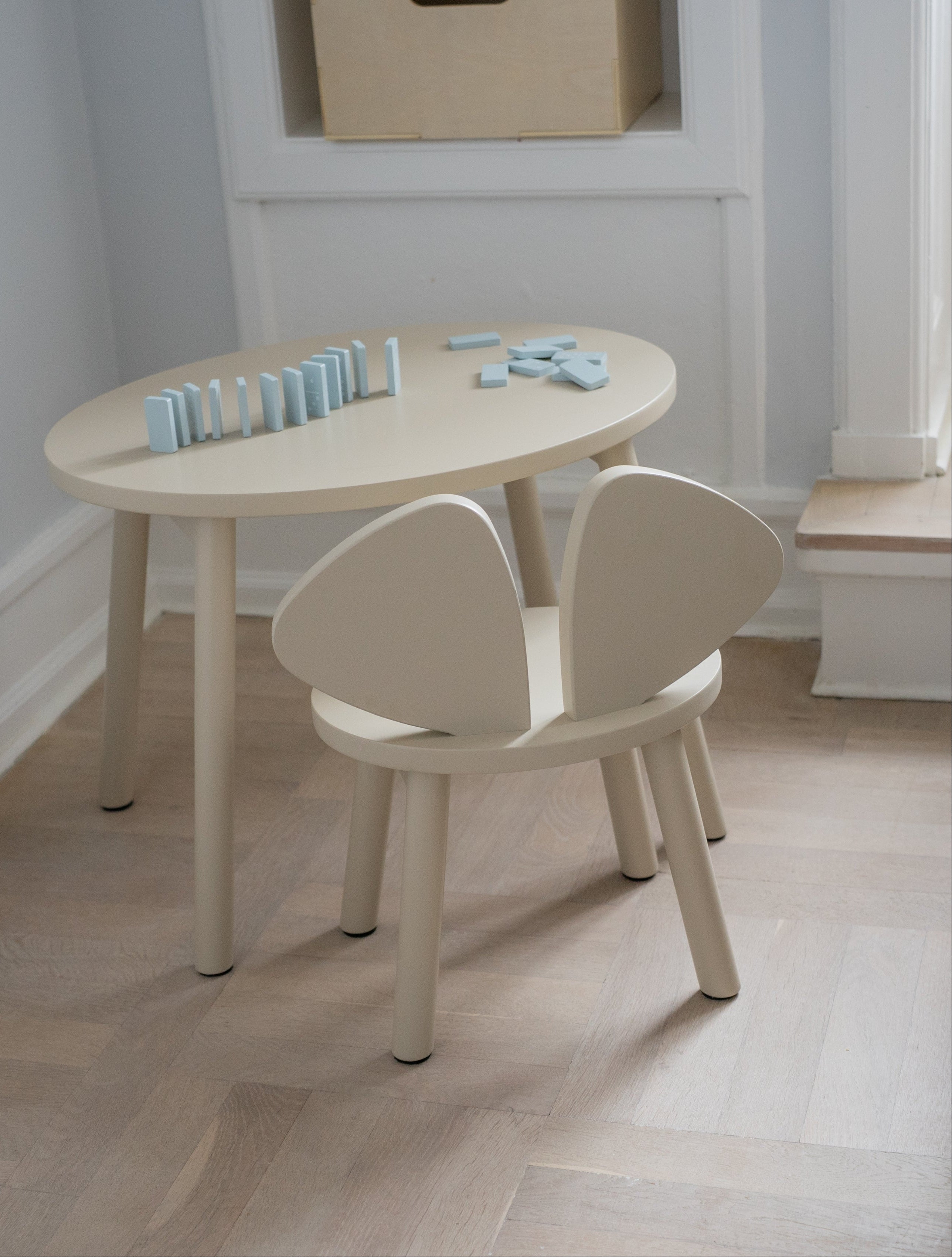 A minimalist children's table and chair set in a light cream or off-white finish, styled in a bright room with a light wood floor and white walls.