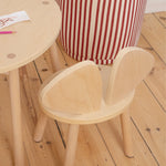 A light natural wood children's chair with a rounded back and matching round wooden table, shown in a room setting with a red and white striped pouf and a wooden floor.