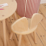 A light natural wood children's chair with a rounded back and matching round wooden table, shown in a room setting with a red and white striped pouf and a wooden floor.