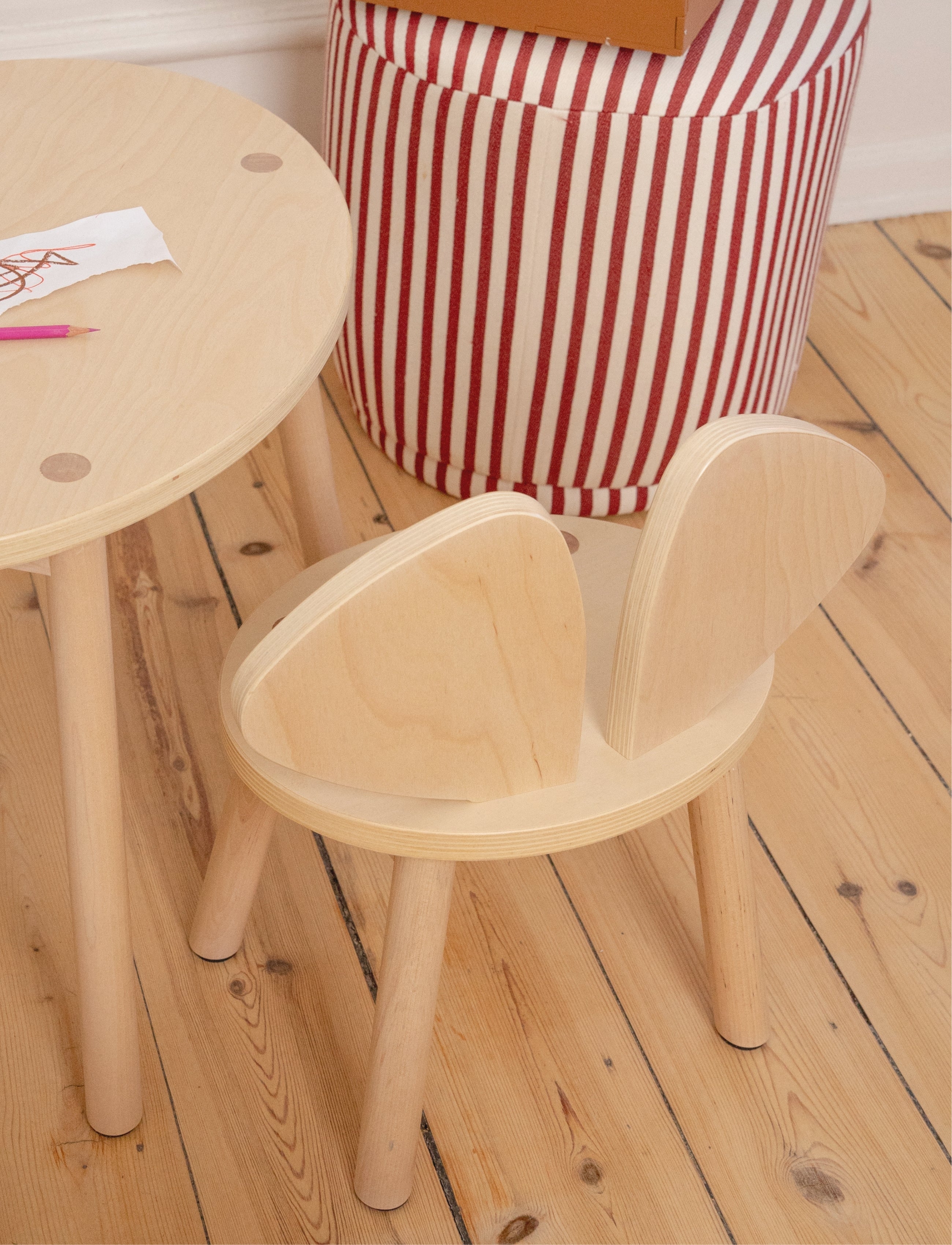 A light natural wood children's chair with a rounded back and matching round wooden table, shown in a room setting with a red and white striped pouf and a wooden floor.