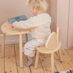A toddler with curly blonde hair sits on a light wood bunny chair at a matching small round wooden table, wearing a cream knit sweater and cream pants. The toddler is holding a blue geometric toy.