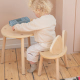 A toddler with curly blonde hair sits on a light wood bunny chair at a matching small round wooden table, wearing a cream knit sweater and cream pants. The toddler is holding a blue geometric toy.