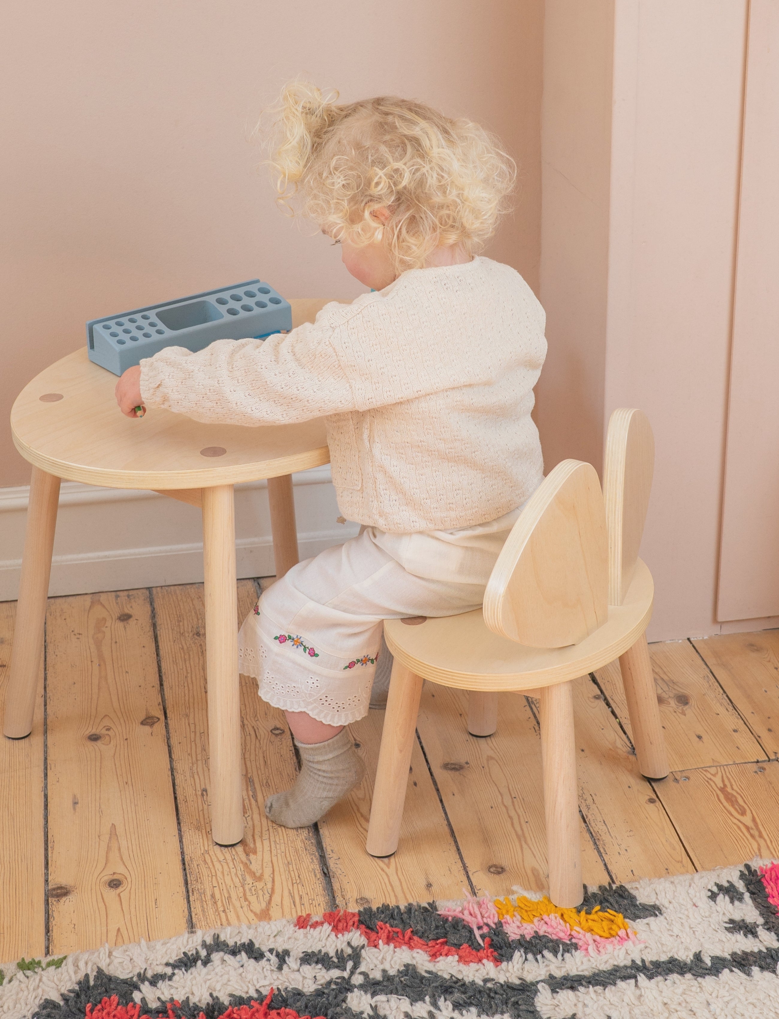 A toddler with curly blonde hair sits on a light wood bunny chair at a matching small round wooden table, wearing a cream knit sweater and cream pants. The toddler is holding a blue geometric toy.