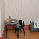 A small black round table and matching black chair in a child's room with patterned wallpaper, wood flooring, and a gray magazine rack.