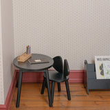 A small black round table and matching black chair in a child's room with patterned wallpaper, wood flooring, and a gray magazine rack.