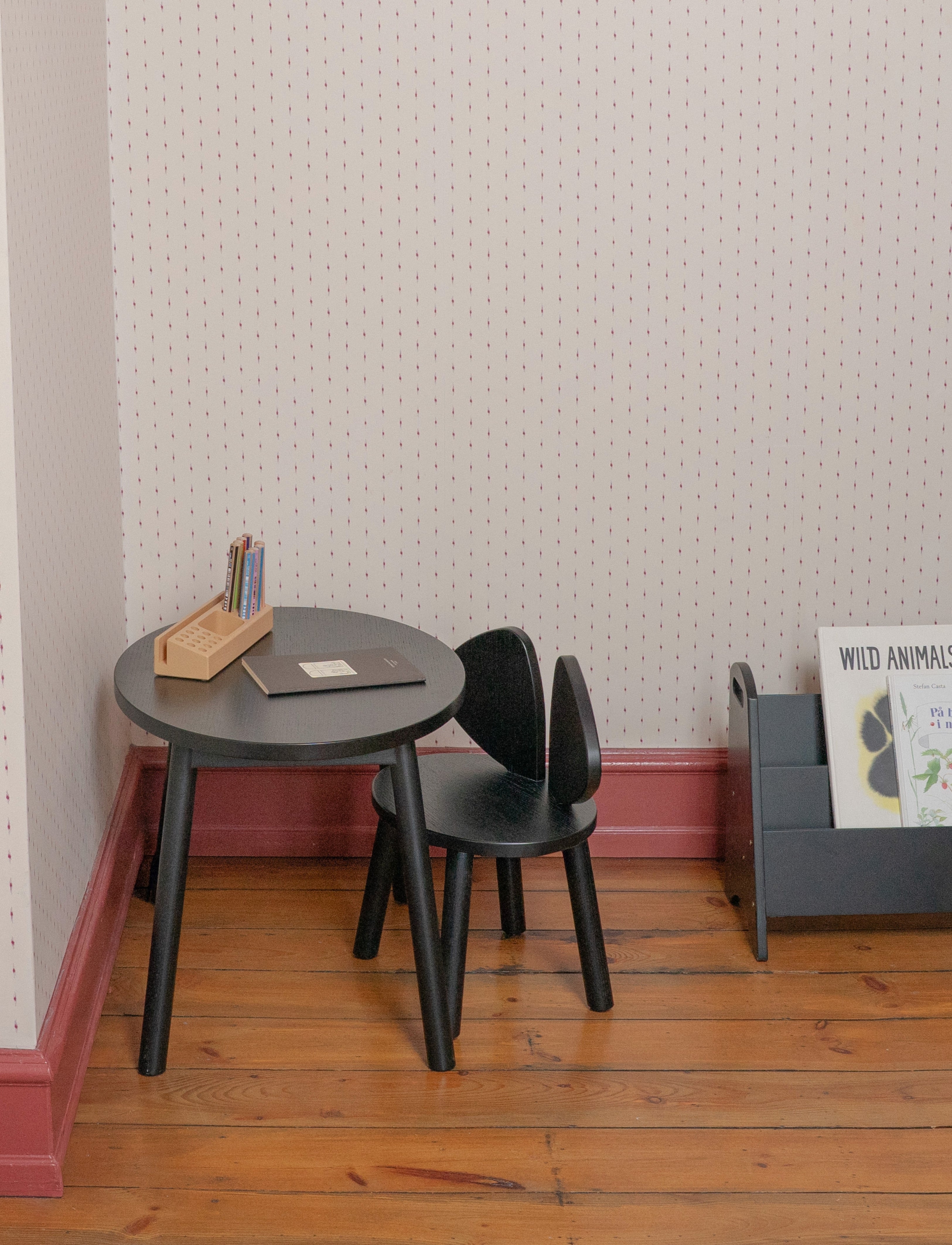 A small black round table and matching black chair in a child's room with patterned wallpaper, wood flooring, and a gray magazine rack.
