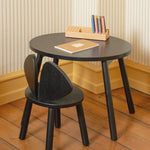 A small round black children's table with three legs and a matching black chair with a heart-shaped backrest, shown in a room with striped wallpaper and a wooden floor. A wooden book holder and a notebook are placed on the table.
