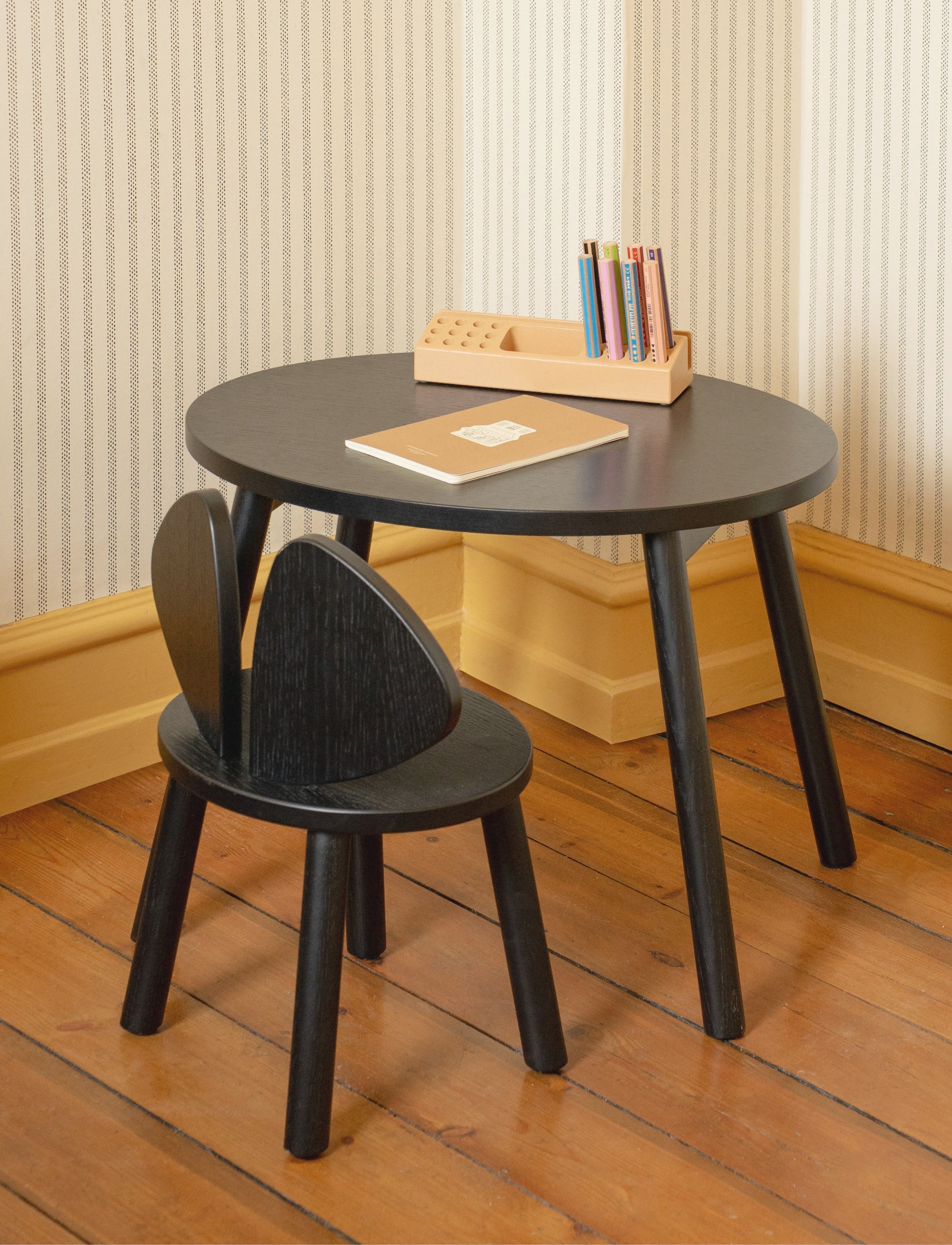 A small round black children's table with three legs and a matching black chair with a heart-shaped backrest, shown in a room with striped wallpaper and a wooden floor. A wooden book holder and a notebook are placed on the table.