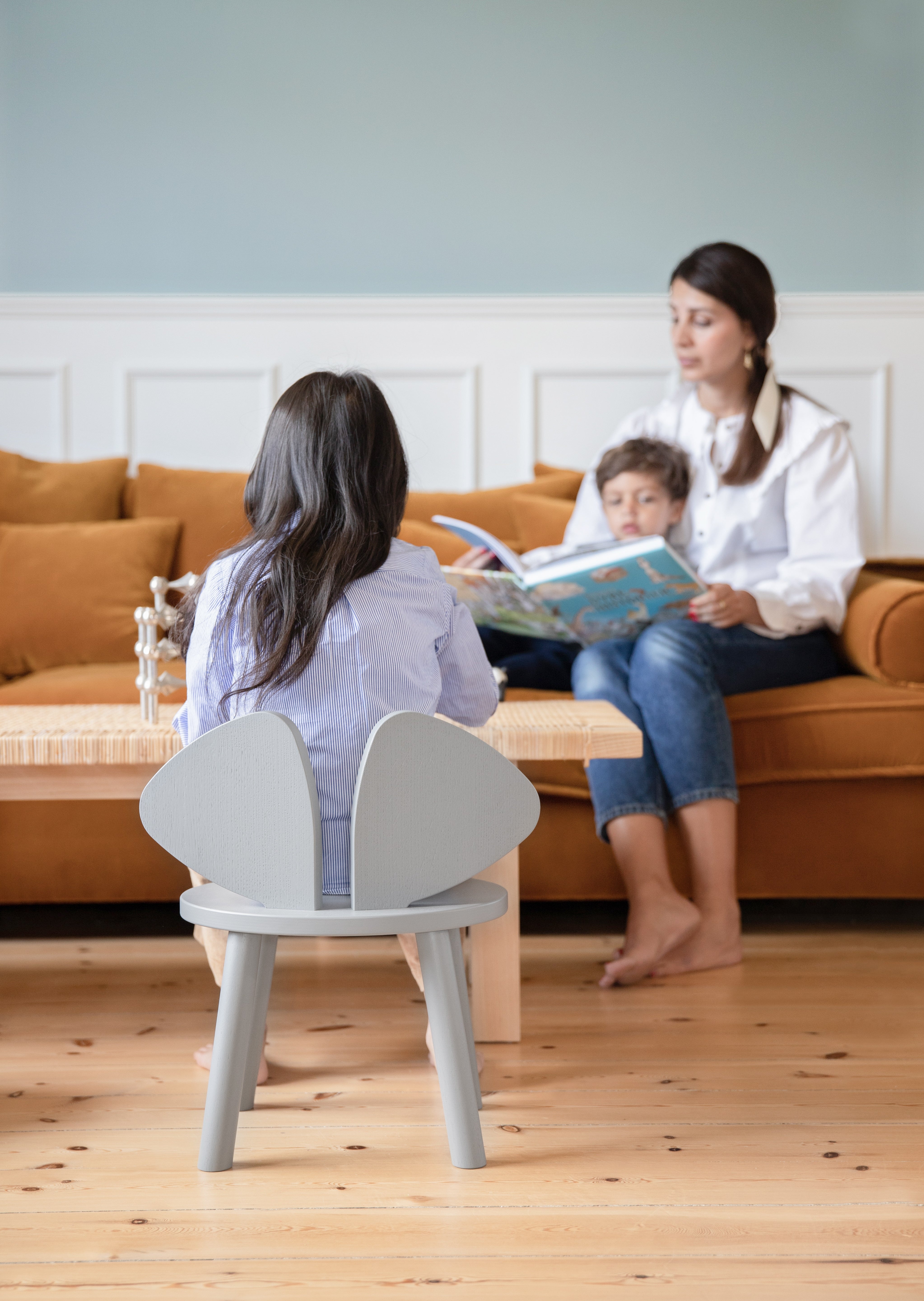 A girl sitting on the grey wooden chair with her mom and brother in front of her
