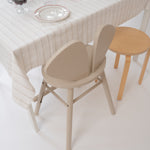 A minimalist dining scene with a cream tablecloth, white plates with red rims, and two chairs: a prominent beige sculptural chair in the foreground and a light wood stool to the right.