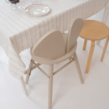 A minimalist dining scene with a cream tablecloth, white plates with red rims, and two chairs: a prominent beige sculptural chair in the foreground and a light wood stool to the right.
