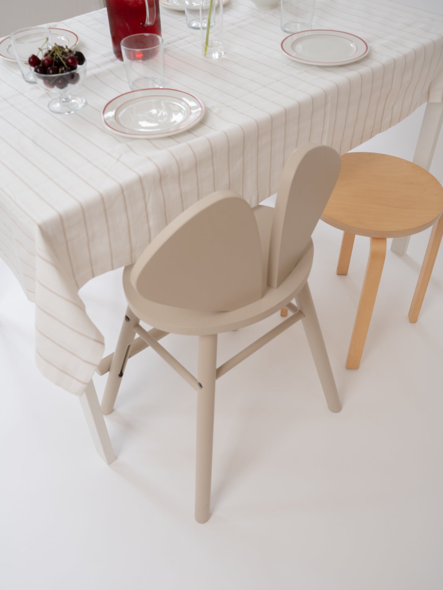 A minimalist dining scene with a cream tablecloth, white plates with red rims, and two chairs: a prominent beige sculptural chair in the foreground and a light wood stool to the right.