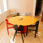 A round dining table with a bright yellow top and red base, surrounded by three chairs in red, black, and beige, set in a sunlit room with light wood floors.