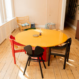 A round dining table with a bright yellow top and red base, surrounded by three chairs in red, black, and beige, set in a sunlit room with light wood floors.