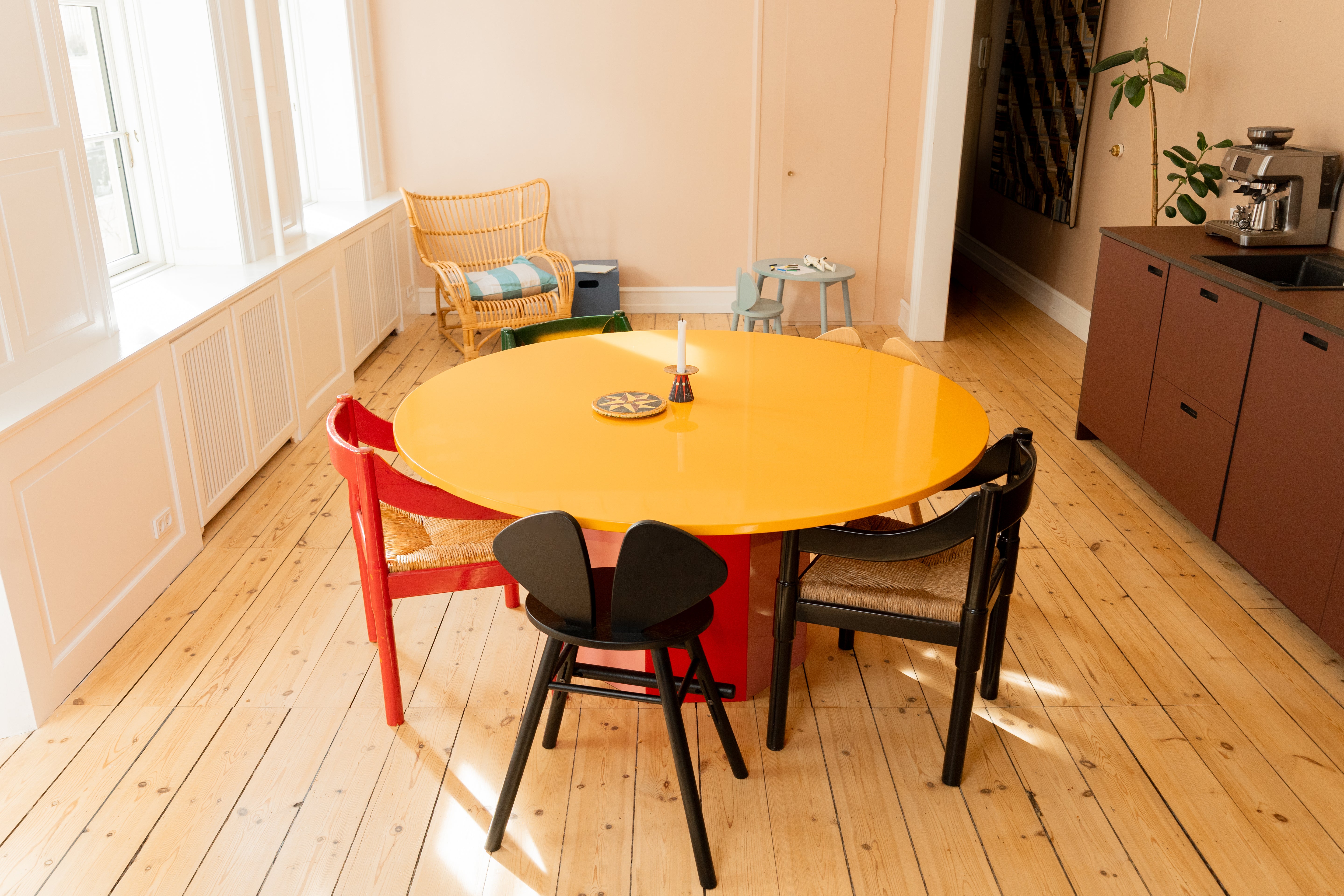 A round dining table with a bright yellow top and red base, surrounded by three chairs in red, black, and beige, set in a sunlit room with light wood floors.