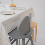 A modern gray wooden dining chair with a curved backrest, shown next to a light wood stool and a table set with a striped tablecloth and plates.