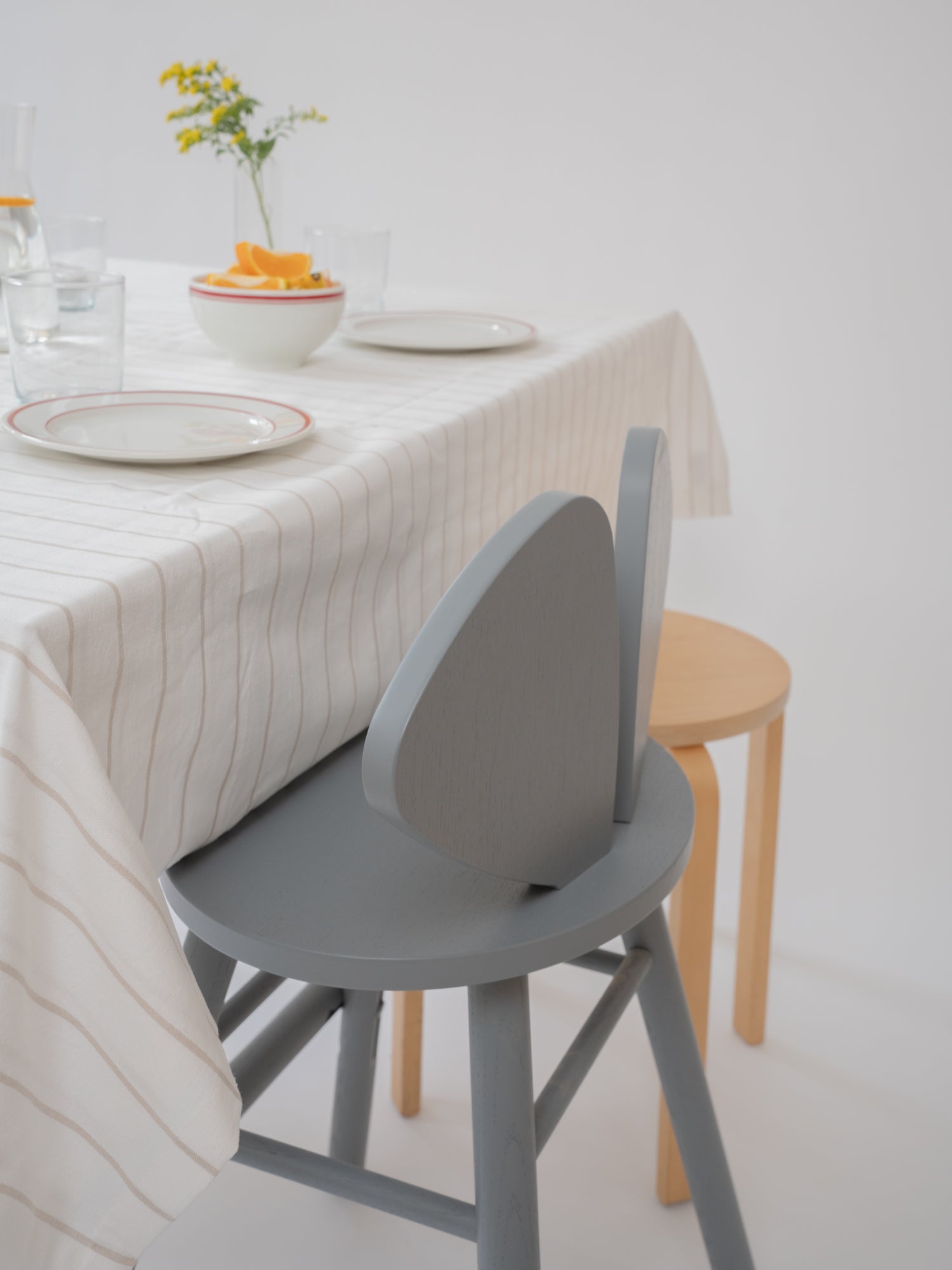 A modern gray wooden dining chair with a curved backrest, shown next to a light wood stool and a table set with a striped tablecloth and plates.