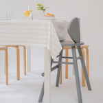 A modern dining setup featuring a gray wooden chair in the foreground, a light wood chair behind it, and a white table with a cream striped tablecloth, set against a clean white background.