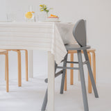 A modern dining setup featuring a gray wooden chair in the foreground, a light wood chair behind it, and a white table with a cream striped tablecloth, set against a clean white background.