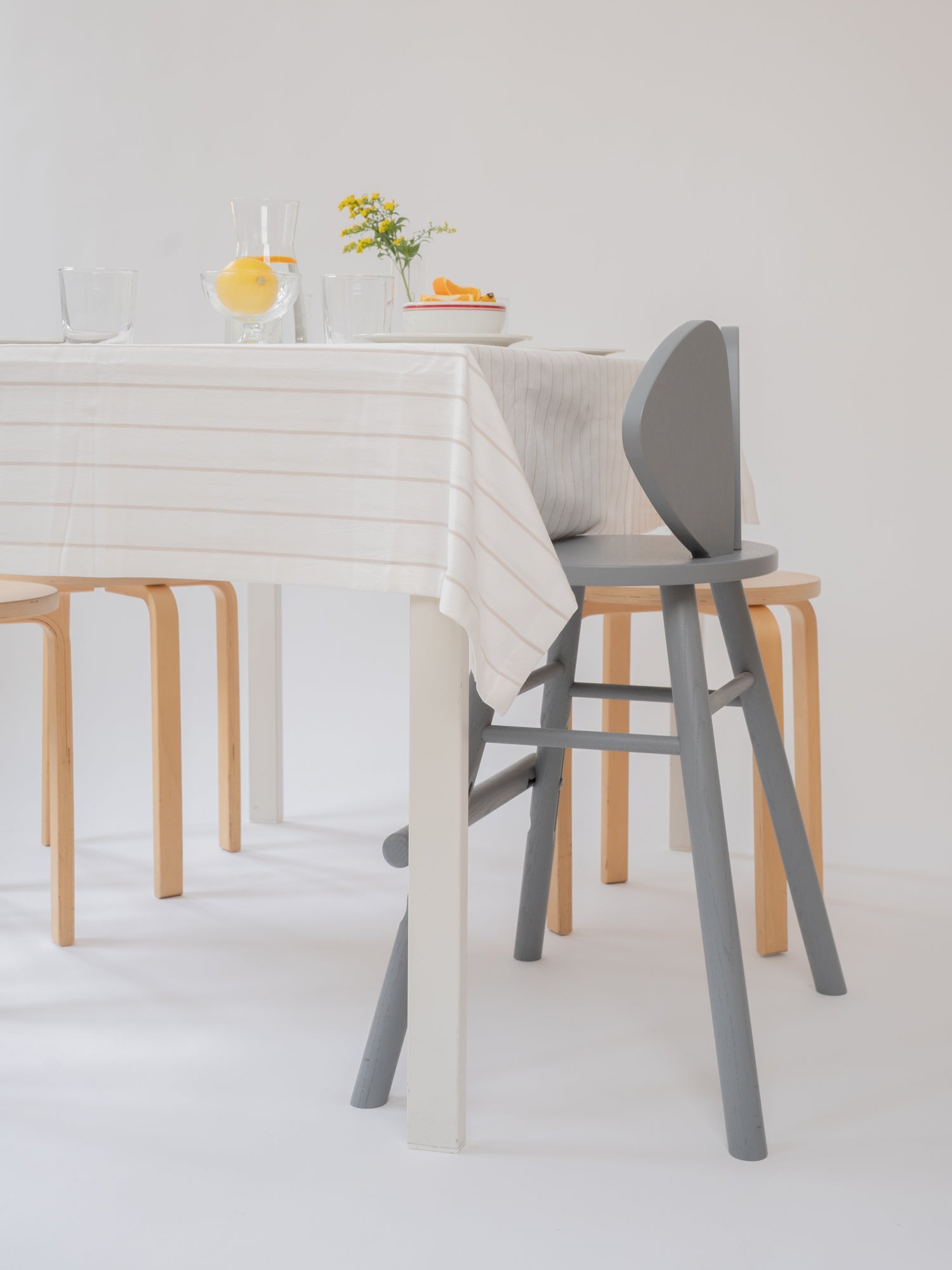 A modern dining setup featuring a gray wooden chair in the foreground, a light wood chair behind it, and a white table with a cream striped tablecloth, set against a clean white background.