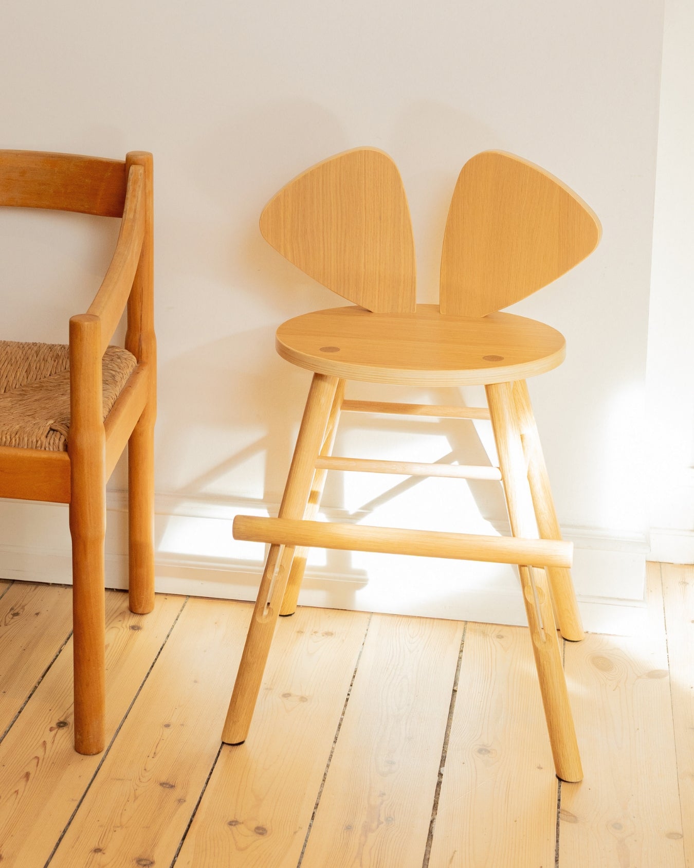 Nofred Mouse Chair Junior in oak beside a woven-seat wooden chair, in sunlight against a white wall.