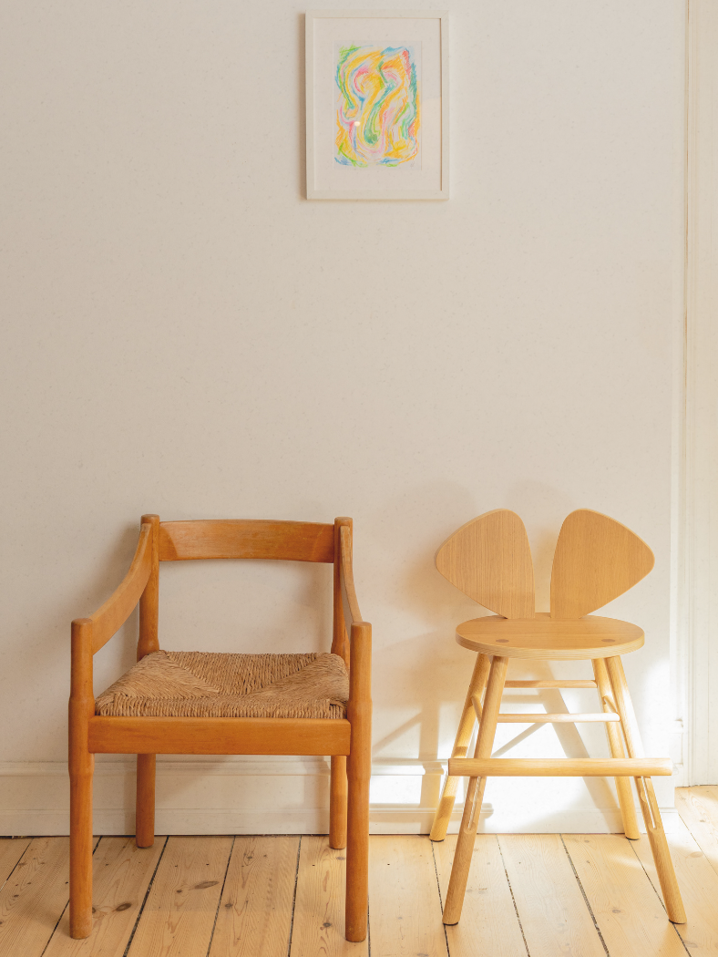 Two wooden chairs, one with a woven seat, against a white wall with a colorful abstract painting.