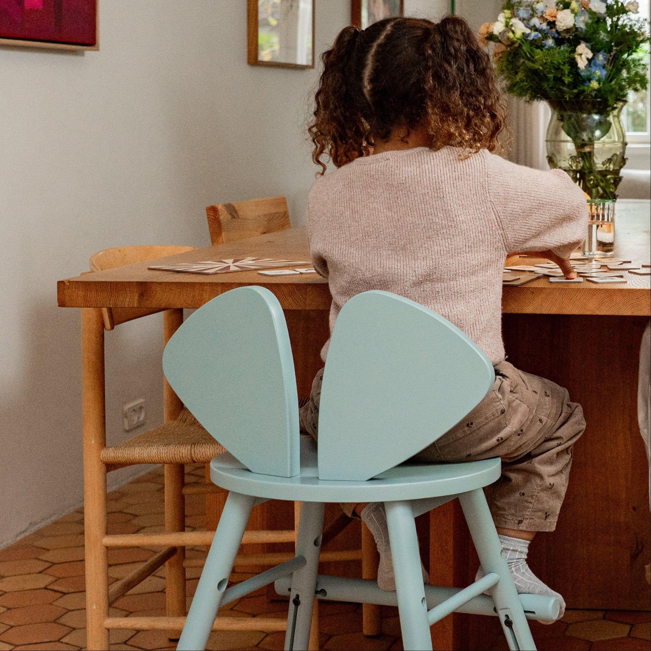 Child sitting at a wooden table with olive green chair in a home setting.