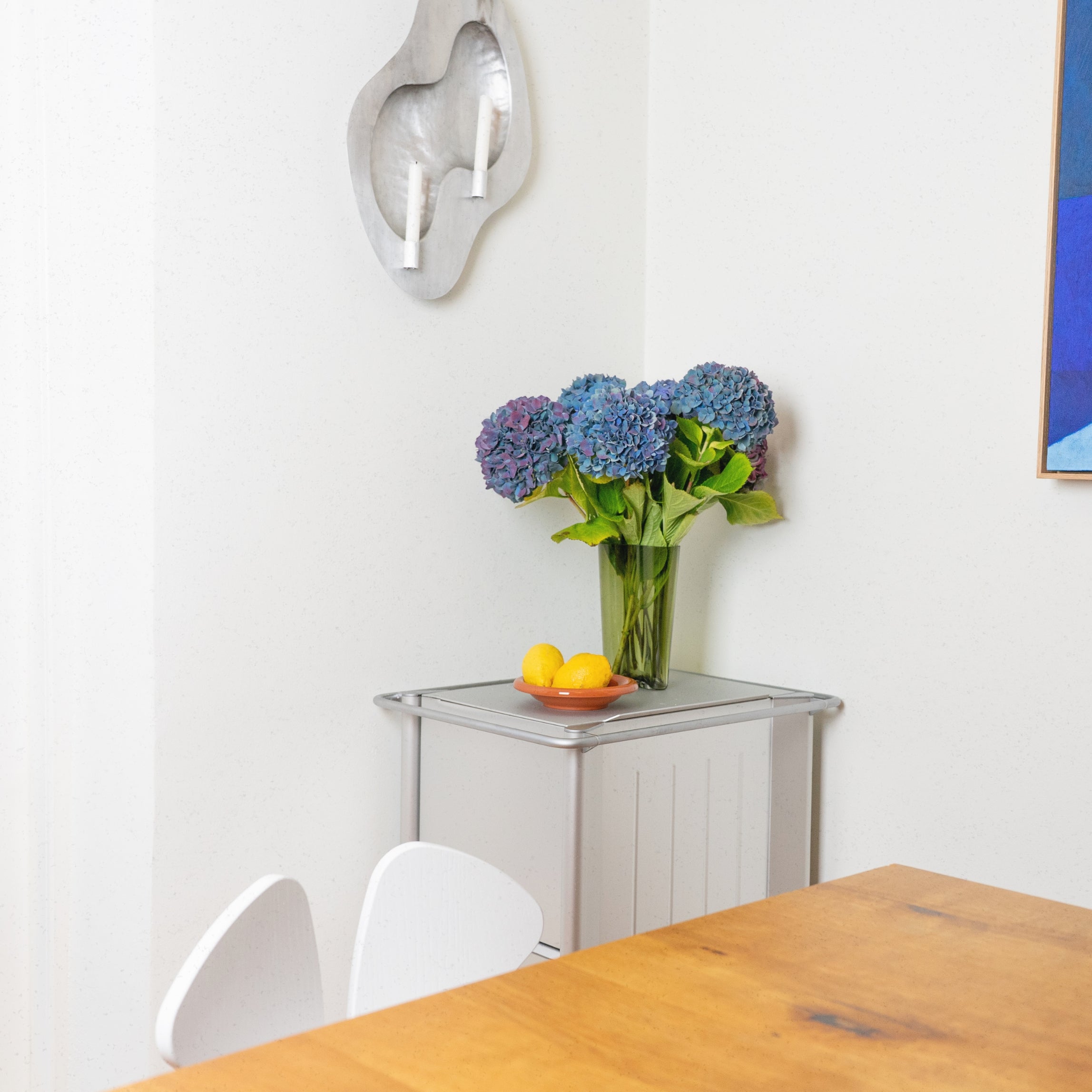 Vase with blue flowers on a glass table in a room with a wooden table and chairs.