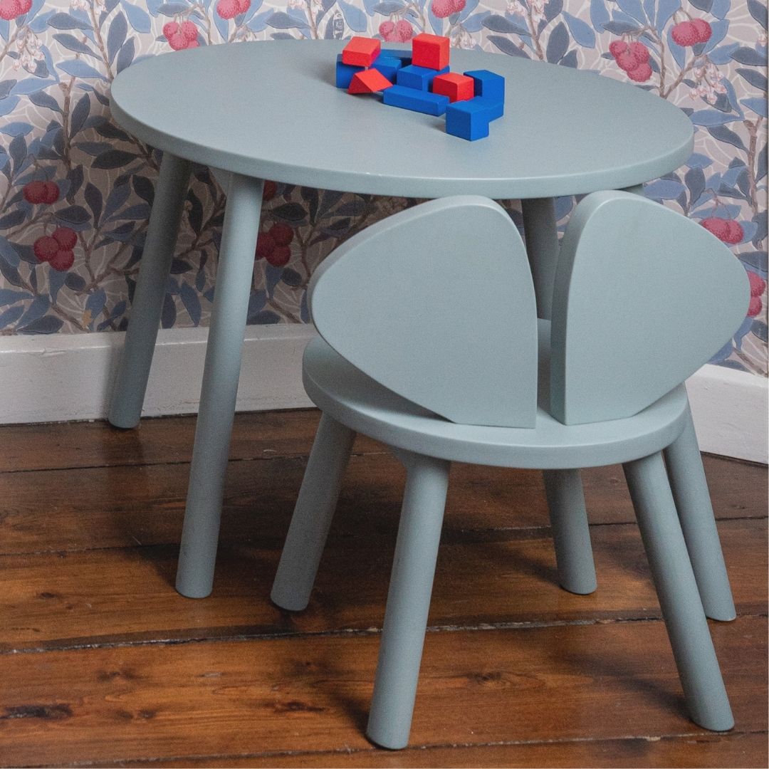 Children's table and chairs set with toys on a wooden floor against a floral wallpapered wall.