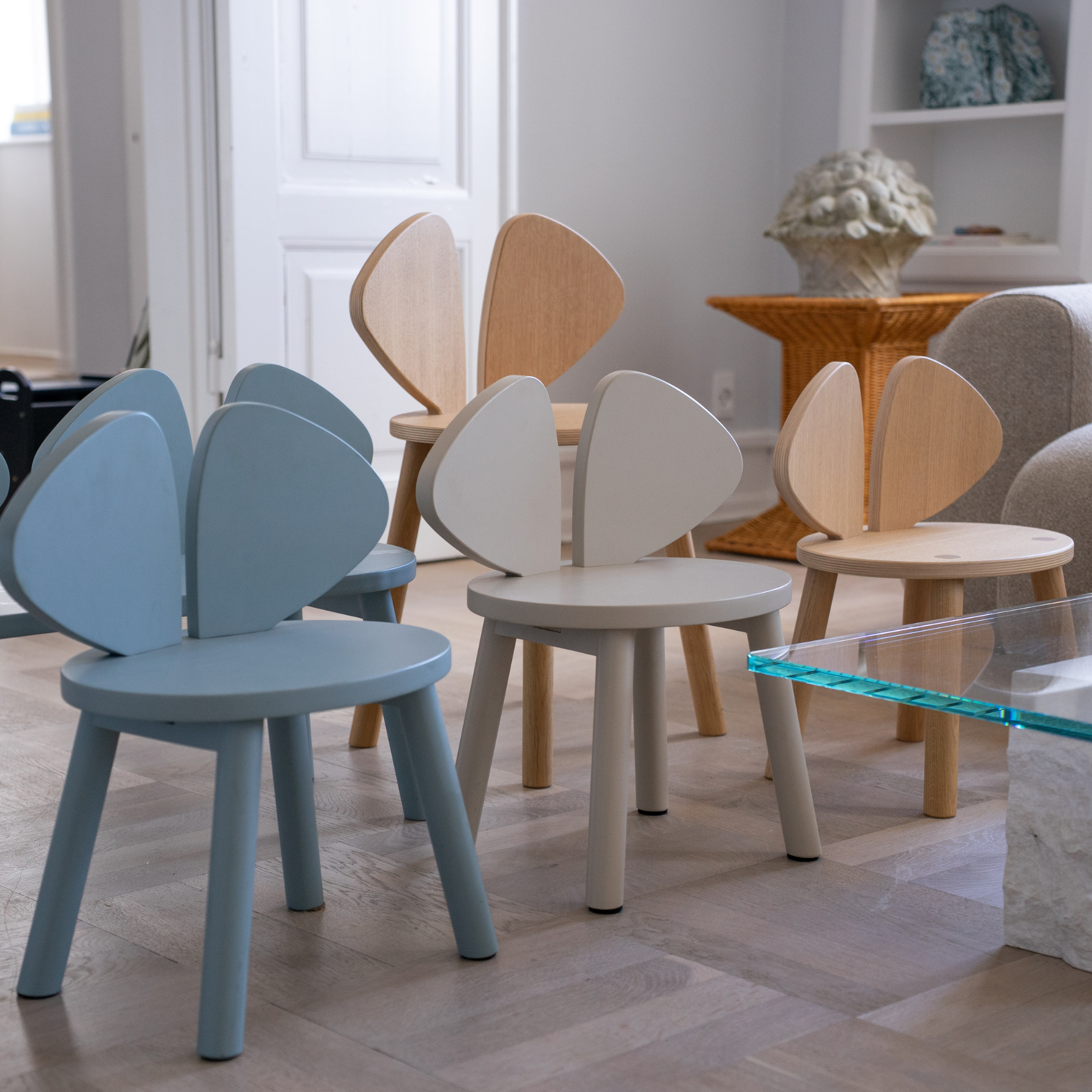 A set of modern wooden children's chairs with curved backrests in light blue, cream, and natural wood finishes, arranged in a bright, contemporary living room.