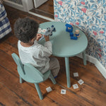Child playing with toys at a green table in a room with wooden floor and floral wallpaper.