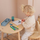Child sitting at a small wooden table with art supplies, drawing on paper.